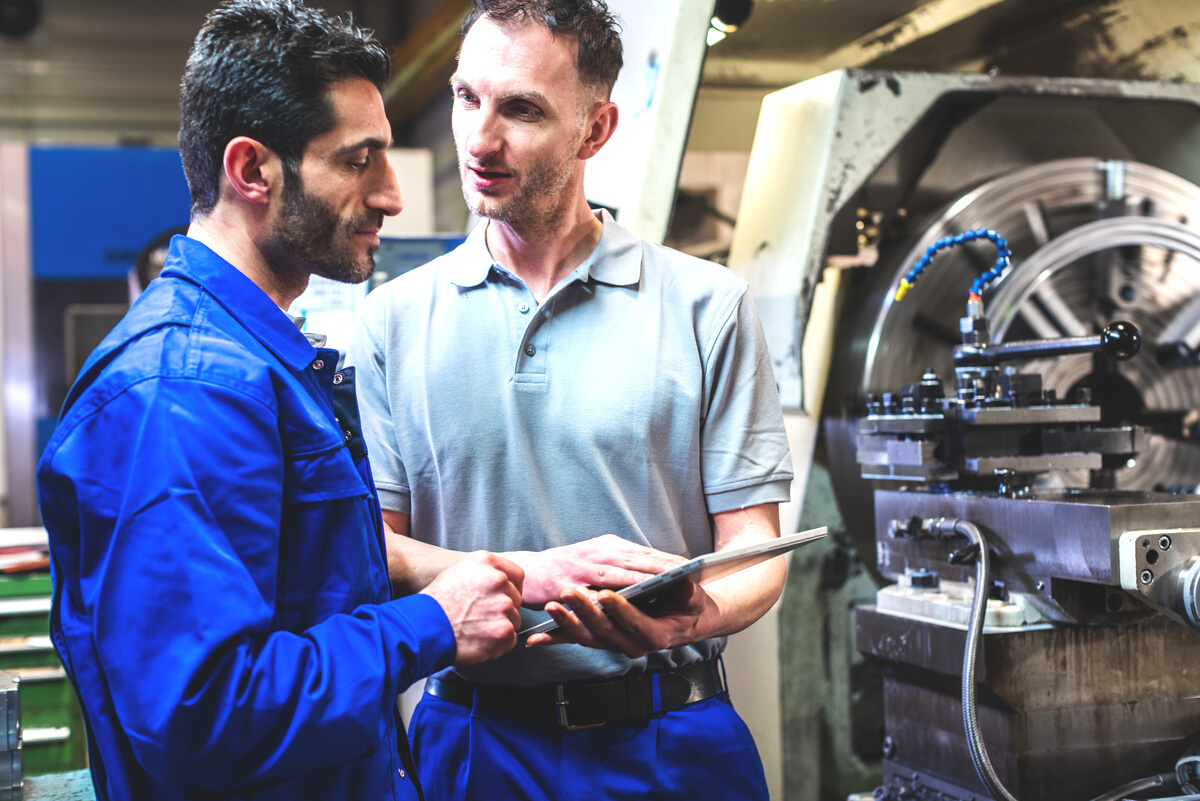 Two Workers Discussing a Project in Front of CNC Lathe Machine